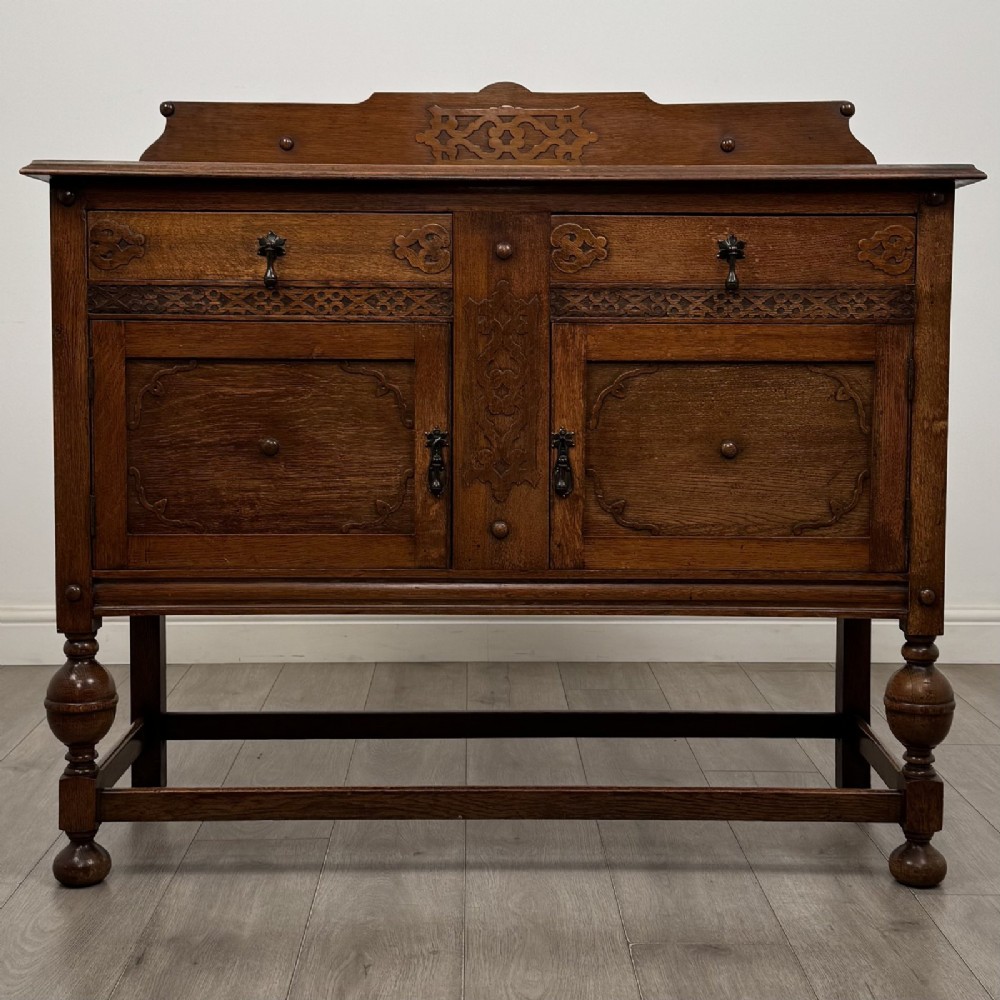 A Jacobean style oak sideboard featuring two drawers, carved door panels, metal drop handles, and turned cup and cover legs connected by low stretchers.