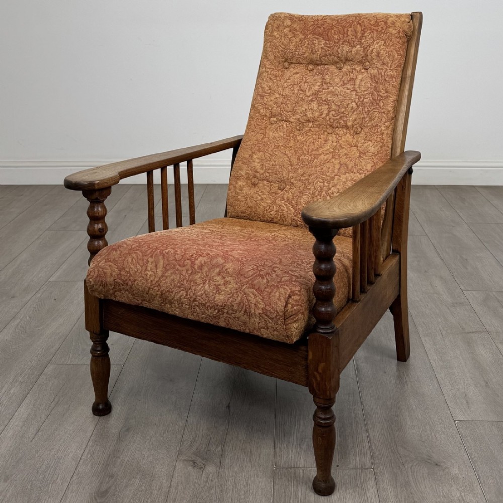 An Arts and Crafts oak reclining armchair with wide armrests, turned front legs, vertical side spindles, and patterned terracotta upholstered cushions.