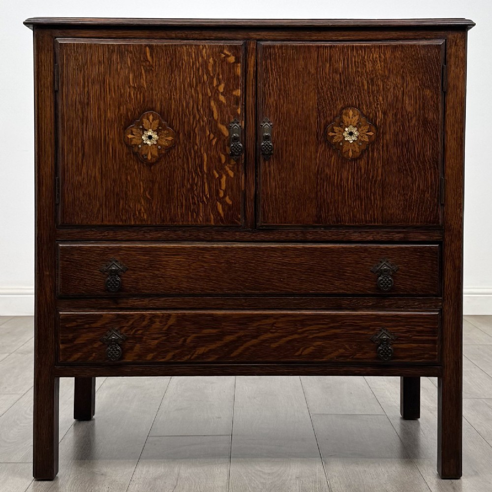 A dark oak Arts and Crafts cupboard featuring double panelled doors with floral marquetry inlays above two wide drawers, finished with dark metal Celtic knot handles.