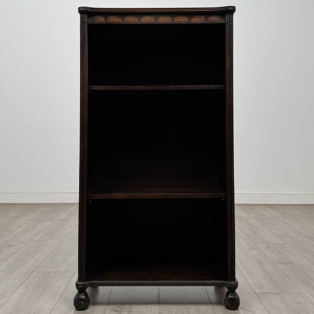 A small dark wood open bookcase featuring three shelves, a carved lunette top frieze, reeded side columns, and turned ball feet.