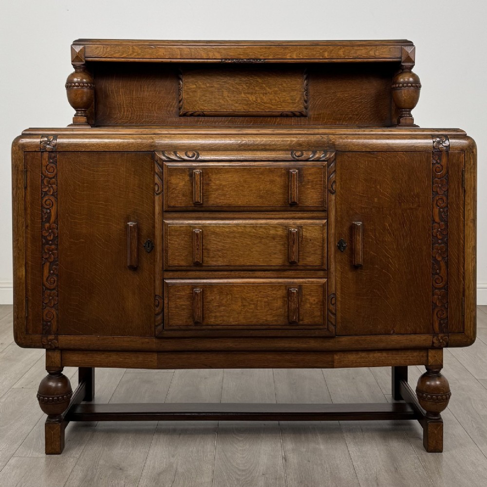 A medium brown oak Jacobean style sideboard featuring a raised gallery shelf, carved side columns, three central drawers, flanking cupboard doors, and bulbous turned legs connected by a stretcher.