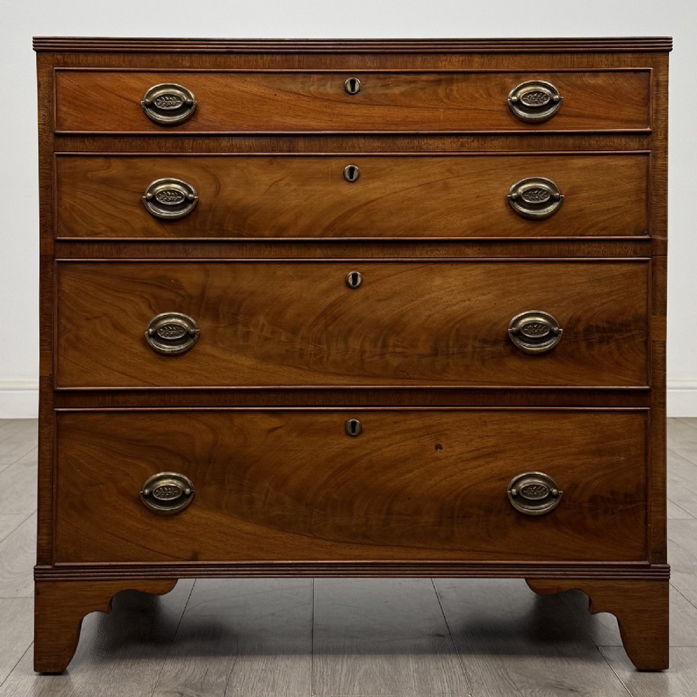 A straight-front Georgian style mahogany chest features four full-width drawers with decorative oval brass drop handles, keyhole escutcheons, and bracket feet.