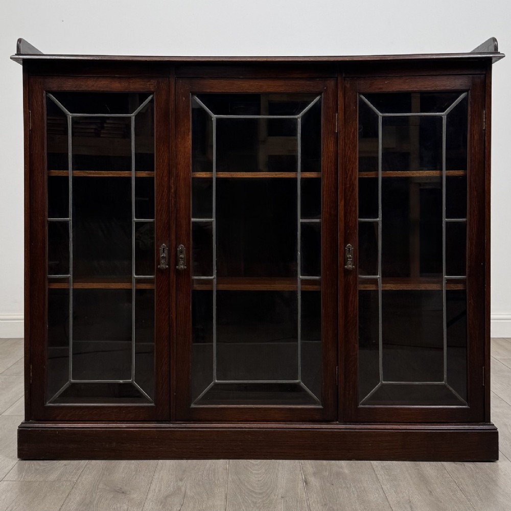 An Edwardian dark oak display cabinet featuring three doors with geometric leaded glass panels and interior wooden shelves.