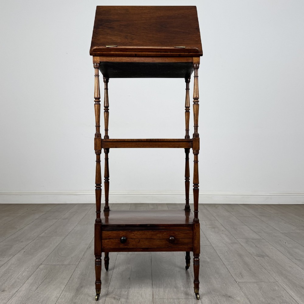 An antique Regency rosewood three-tiered whatnot featuring an adjustable angled reading slope top, slender turned upright supports, a bottom drawer, and brass castors.