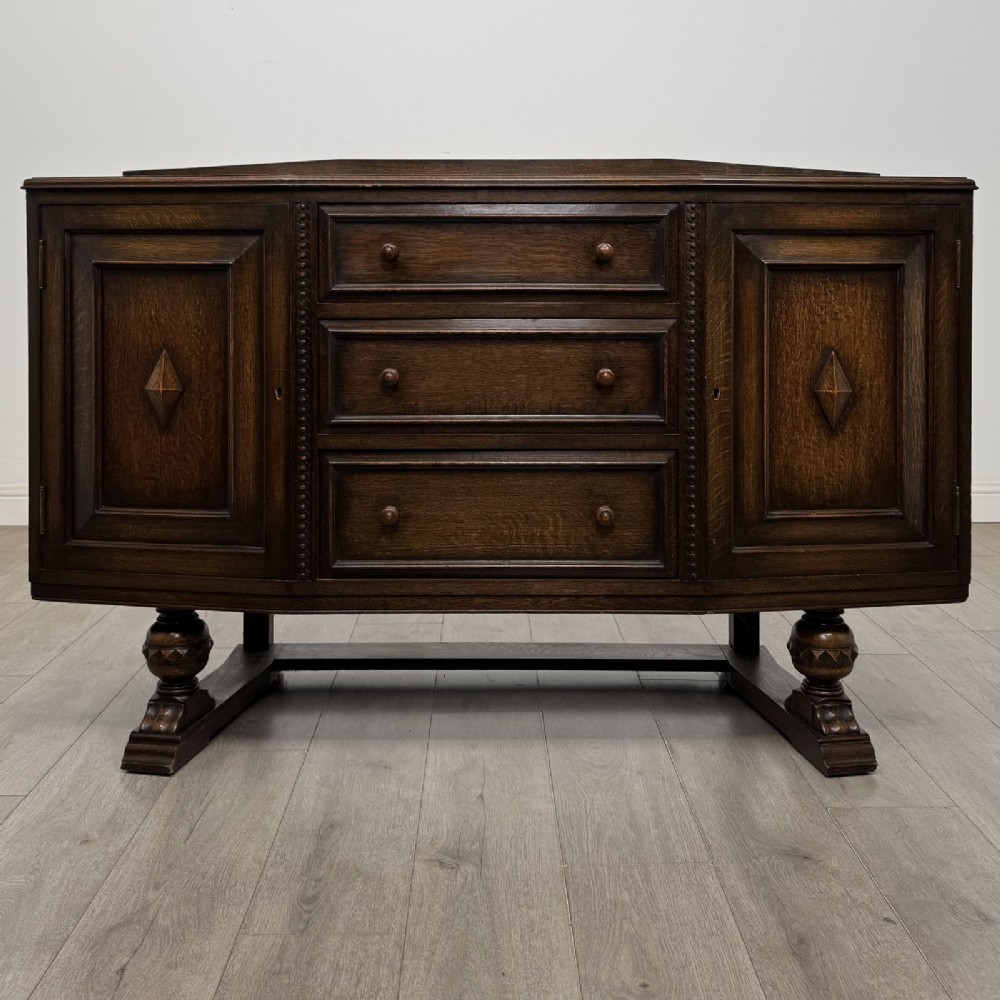 A dark brown Jacobean style oak sideboard featuring three central drawers flanked by two geometric panel cupboard doors, supported on turned legs.
