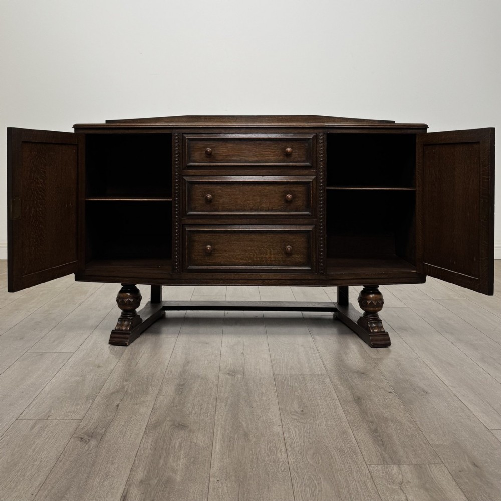 A dark brown Jacobean style oak sideboard featuring three central drawers flanked by two geometric panel cupboard doors, supported on turned legs. View II