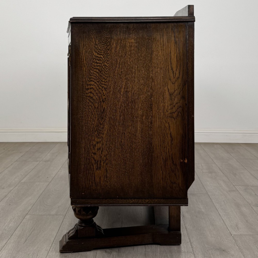 A dark brown Jacobean style oak sideboard featuring three central drawers flanked by two geometric panel cupboard doors, supported on turned legs. View IV