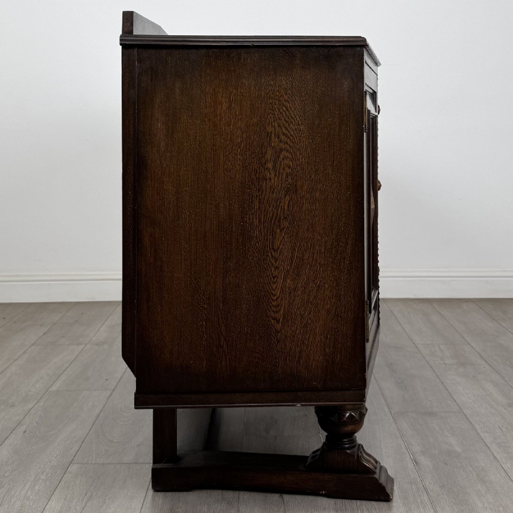 A dark brown Jacobean style oak sideboard featuring three central drawers flanked by two geometric panel cupboard doors, supported on turned legs. View VIII
