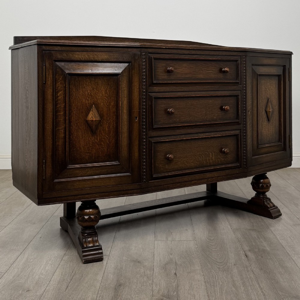 A dark brown Jacobean style oak sideboard featuring three central drawers flanked by two geometric panel cupboard doors, supported on turned legs. View IX