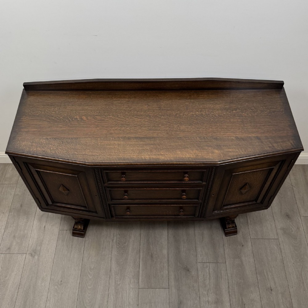A dark brown Jacobean style oak sideboard featuring three central drawers flanked by two geometric panel cupboard doors, supported on turned legs. View X