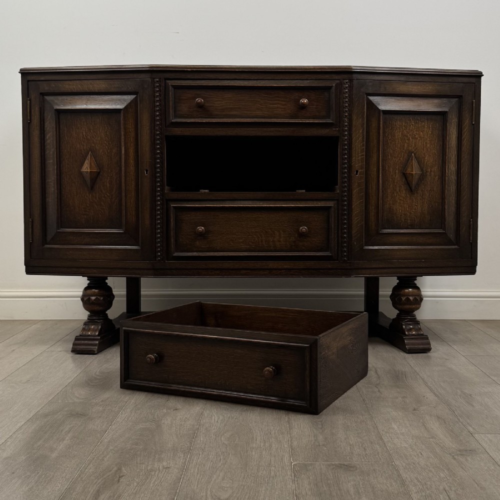 A dark brown Jacobean style oak sideboard featuring three central drawers flanked by two geometric panel cupboard doors, supported on turned legs. View XI