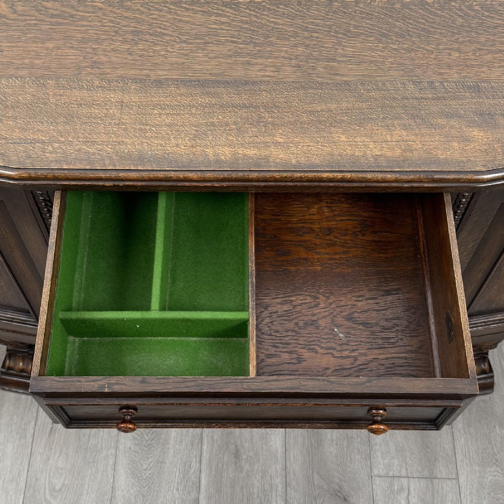 A dark brown Jacobean style oak sideboard featuring three central drawers flanked by two geometric panel cupboard doors, supported on turned legs. View XIII