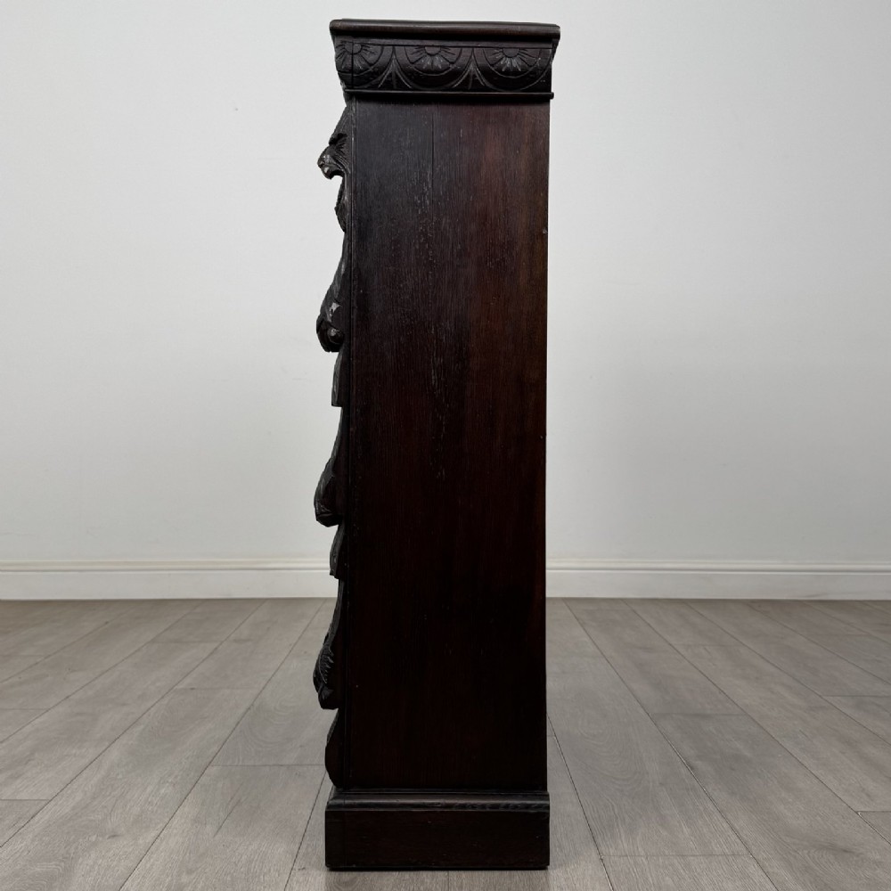 A dark carved oak open bookcase featuring three shelf surfaces and pilasters detailed with foliate carvings and Green Man masks. View IV