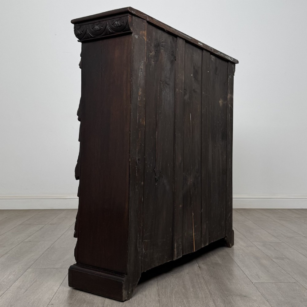 A dark carved oak open bookcase featuring three shelf surfaces and pilasters detailed with foliate carvings and Green Man masks. View V