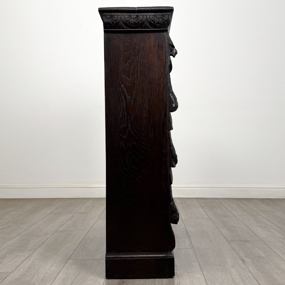 A dark carved oak open bookcase featuring three shelf surfaces and pilasters detailed with foliate carvings and Green Man masks. View VIII