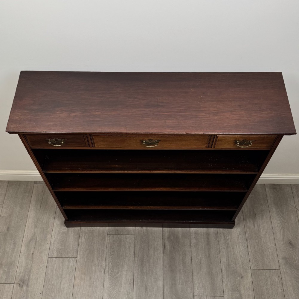 An Edwardian dark mahogany open bookcase featuring three frieze drawers with brass drop handles and three adjustable wooden shelves above a plinth base. View X