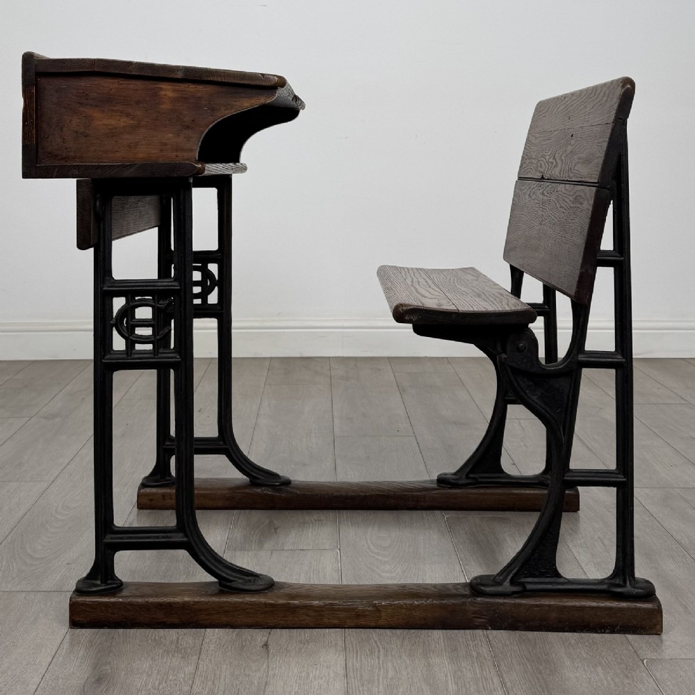 An antique early 20th-century school desk featuring a sloped dark timber top with a storage compartment, attached to a black cast iron frame with an integrated bench seat. View II