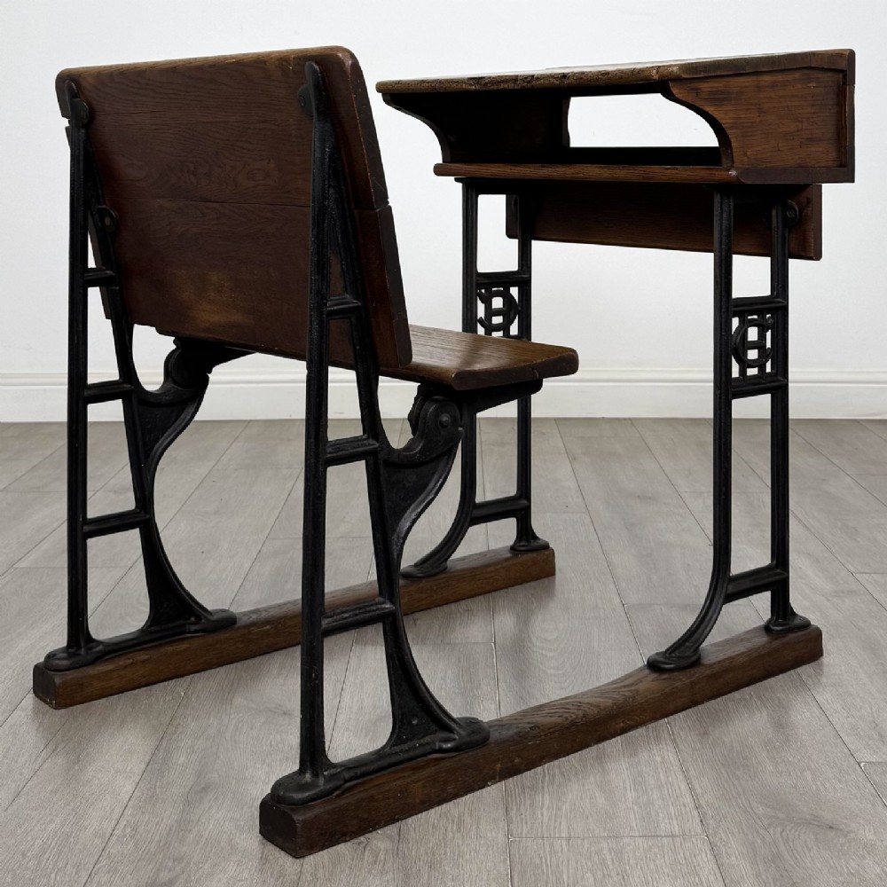 An antique early 20th-century school desk featuring a sloped dark timber top with a storage compartment, attached to a black cast iron frame with an integrated bench seat. View V