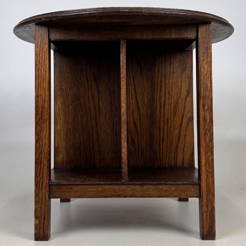 An antique dark oak Arts and Crafts side table featuring a circular top, four square legs, and an integrated four-compartment book rack beneath. View II