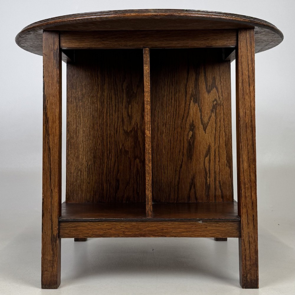 An antique dark oak Arts and Crafts side table featuring a circular top, four square legs, and an integrated four-compartment book rack beneath. View IV