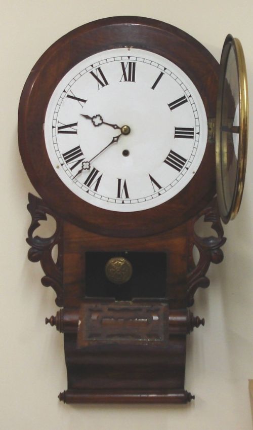 fig 1. An antique American Ansonia 8-day drop-dial wall clock featuring a mahogany wooden case, white dial with Roman numerals, and carved lower scrollwork.