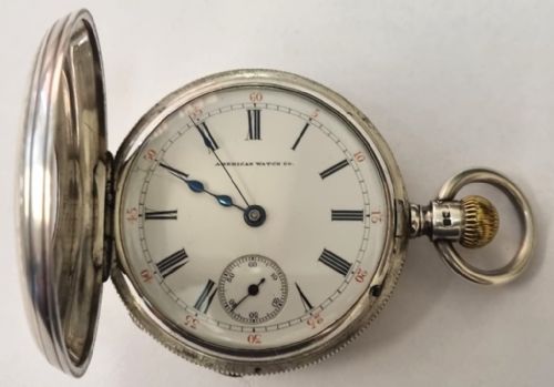 fig 2. A silver half-hunter pocket watch with blue enamel Roman numerals surrounding a central circular glass window revealing dark watch hands.