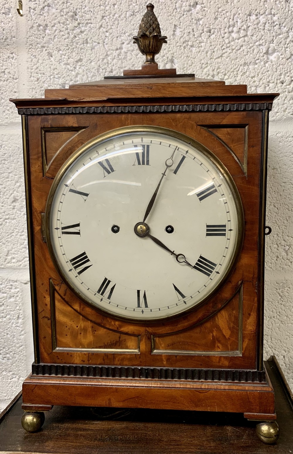 An antique Regency flame mahogany twin fusee bracket clock featuring a round white dial with Roman numerals, a brass pineapple finial, brass inlay, reeded borders, and brass ball feet.