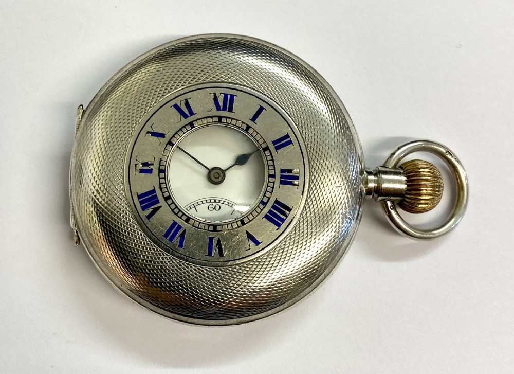 An antique sterling silver half hunter pocket watch featuring an engine-turned textured case and blue enamel Roman numerals around the central aperture.