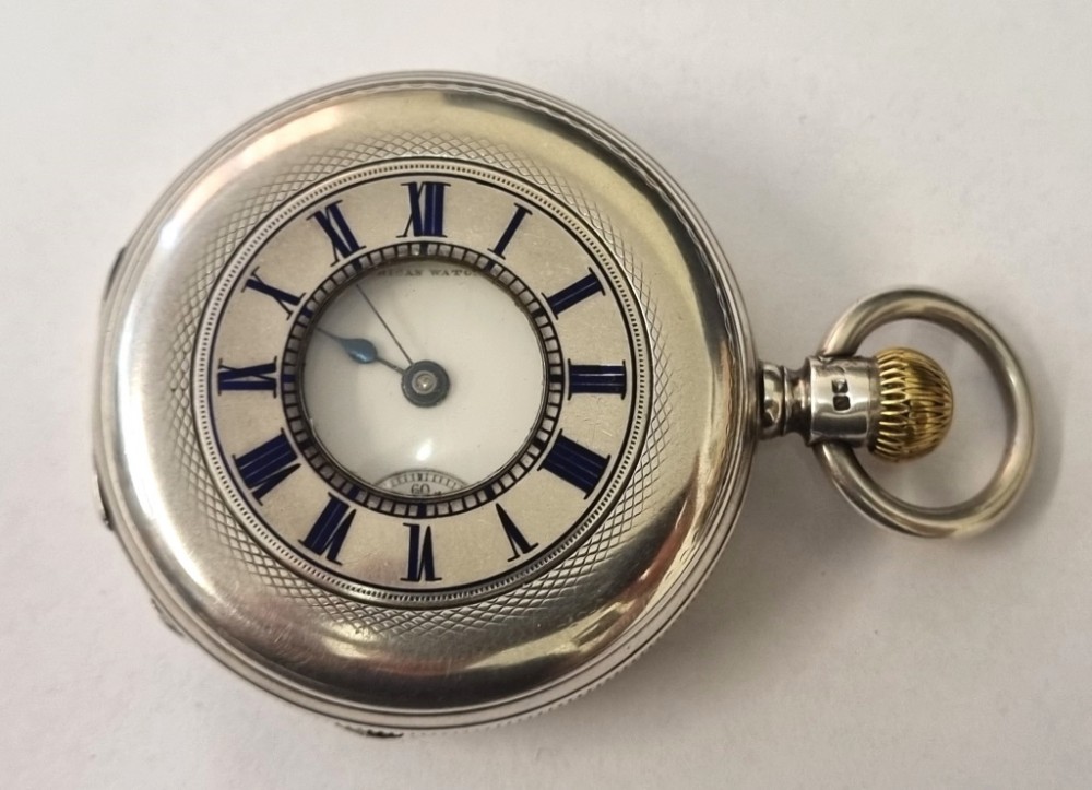 A silver half-hunter pocket watch with blue enamel Roman numerals surrounding a central circular glass window revealing dark watch hands.