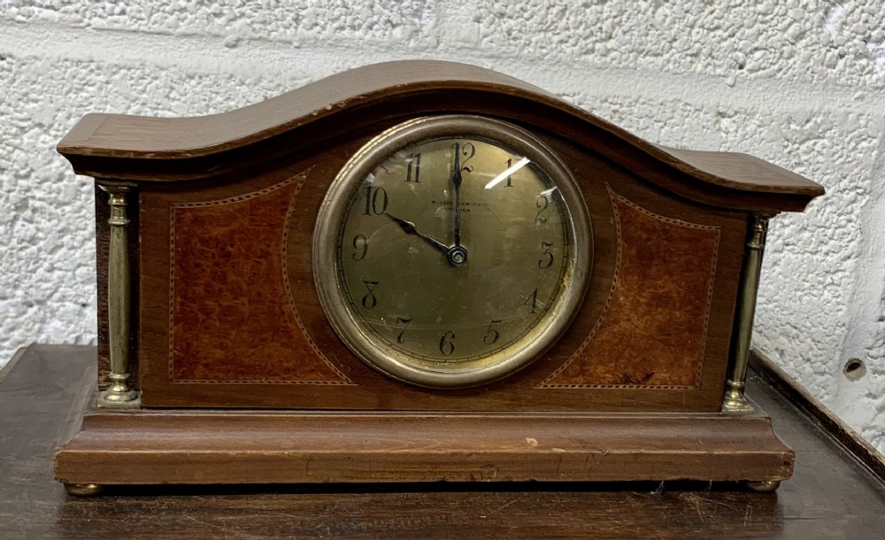 An Edwardian mahogany and walnut veneer arched mantel clock featuring brass side columns, burr wood inlay panels, and a round brass bezel with Arabic numerals.