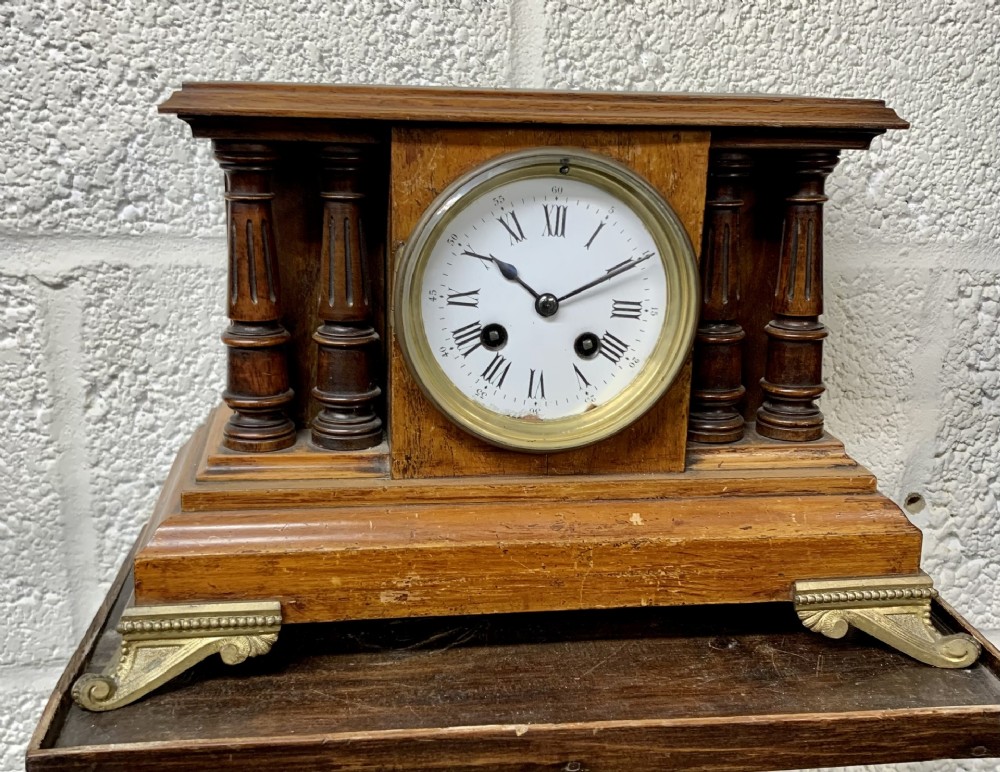 A wooden mantel clock featuring twin turned columns, a white circular dial with Roman numerals, and decorative brass feet.