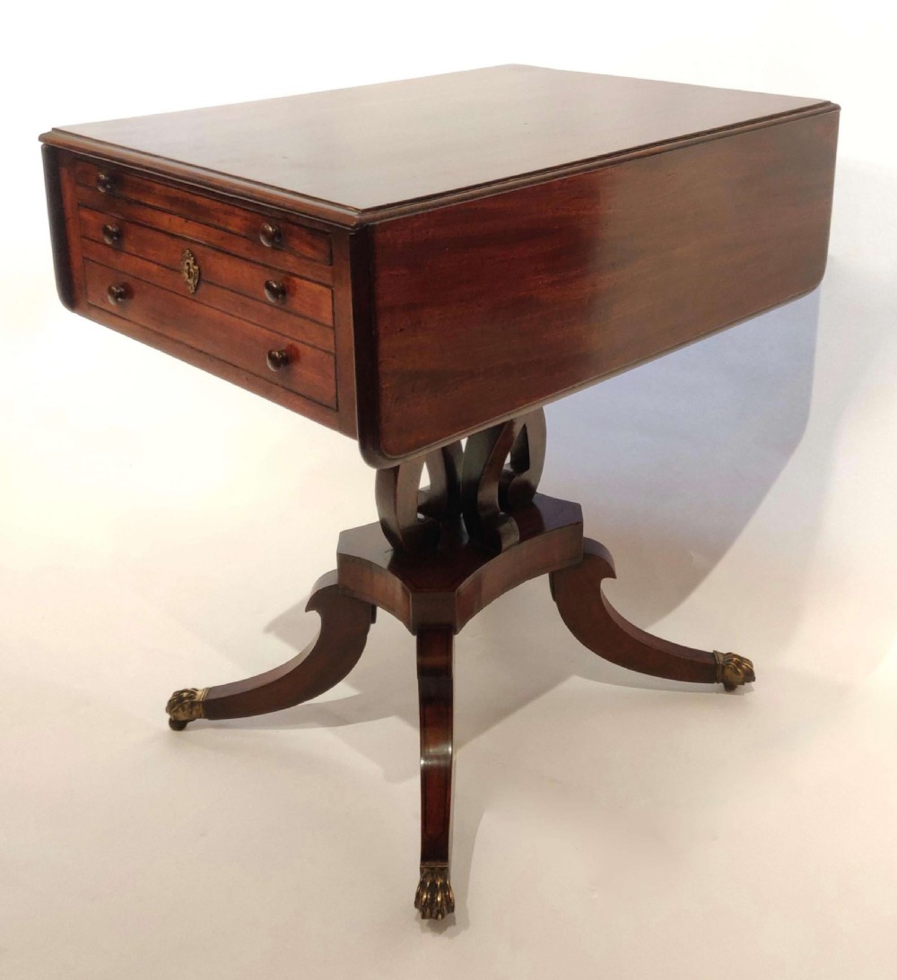 A Regency mahogany drop flap table featuring two drawers, a central carved pedestal, and four splayed legs with brass lion paw castors.