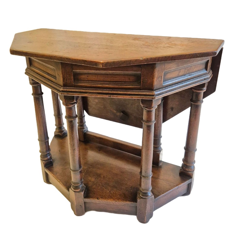 A dark oak credence side table featuring a canted top, turned column legs, a lower shelf, and a fold-over leaf mechanism.