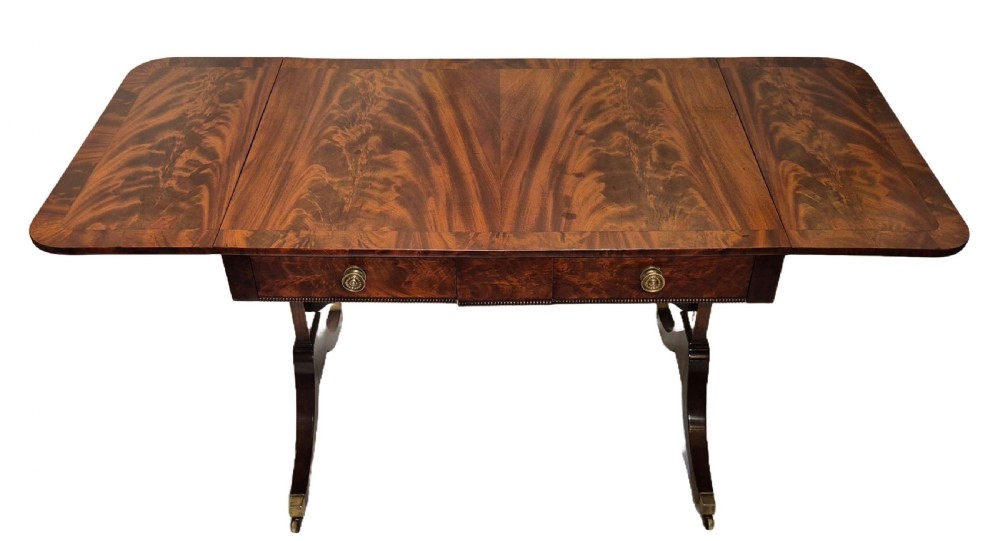 A Regency mahogany sofa table with flame mahogany veneers, twin drop leaves, two frieze drawers with brass handles, and sweeping legs on brass castors.