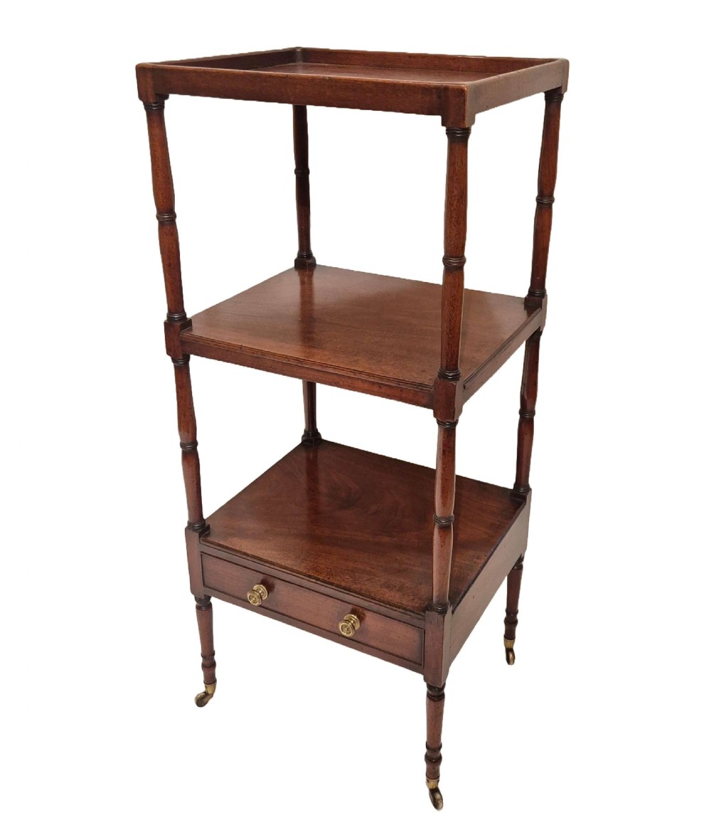 A Georgian mahogany three-tier whatnot featuring turned upright supports, a gallery-topped upper shelf, a single lower drawer with brass knobs, and brass castors.