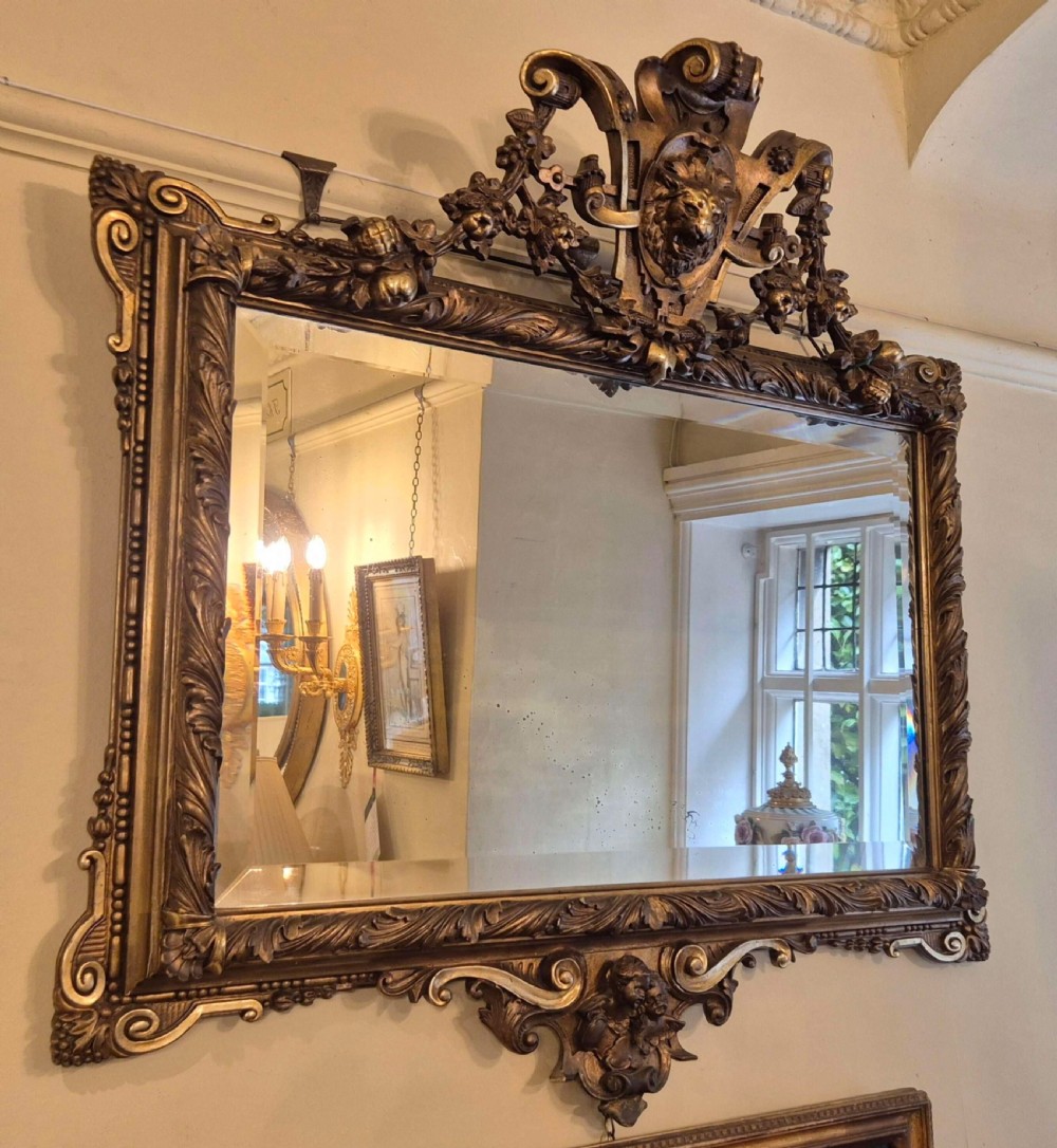 A rectangular giltwood and gesso wall mirror featuring an ornate pediment sculpted with a lion head mask and acanthus leaf details.
