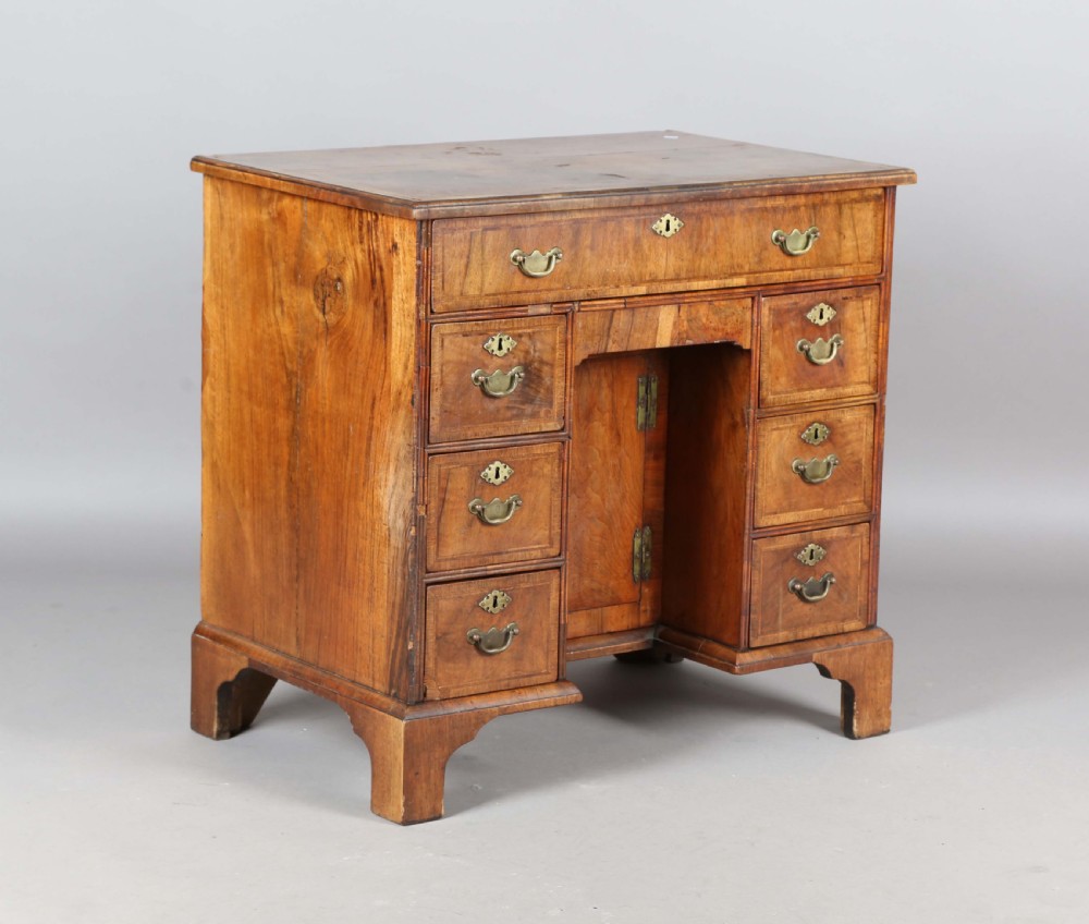 An antique Queen Anne walnut kneehole desk featuring a long top drawer above six side drawers, a central recessed cupboard, and brass drop handles, supported by bracket feet.