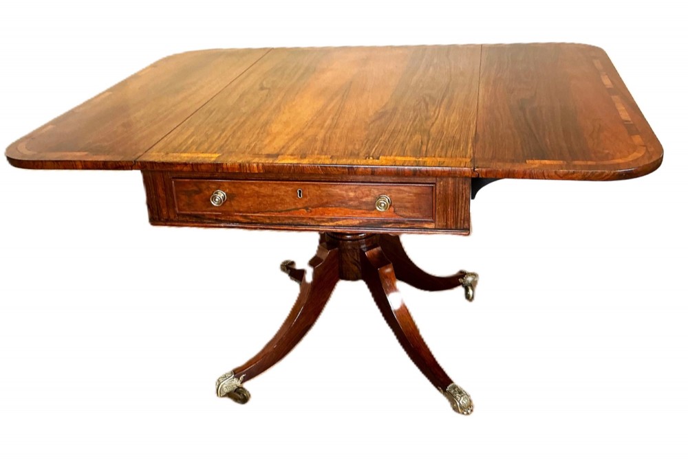 A Regency period rosewood drop-flap library table with a crossbanded top, central drawer, and quadform splayed legs with brass castors.