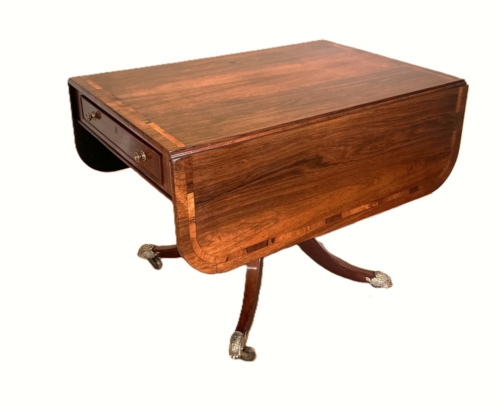 A Regency period rosewood drop-flap library table with a crossbanded top, central drawer, and quadform splayed legs with brass castors. View II