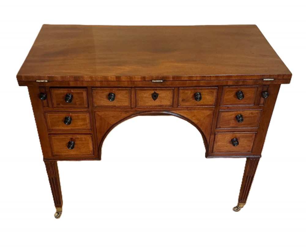 An antique George III mahogany library table featuring an arched central aperture, multiple drawers with dark turned knobs, and reeded tapered legs on brass castors.
