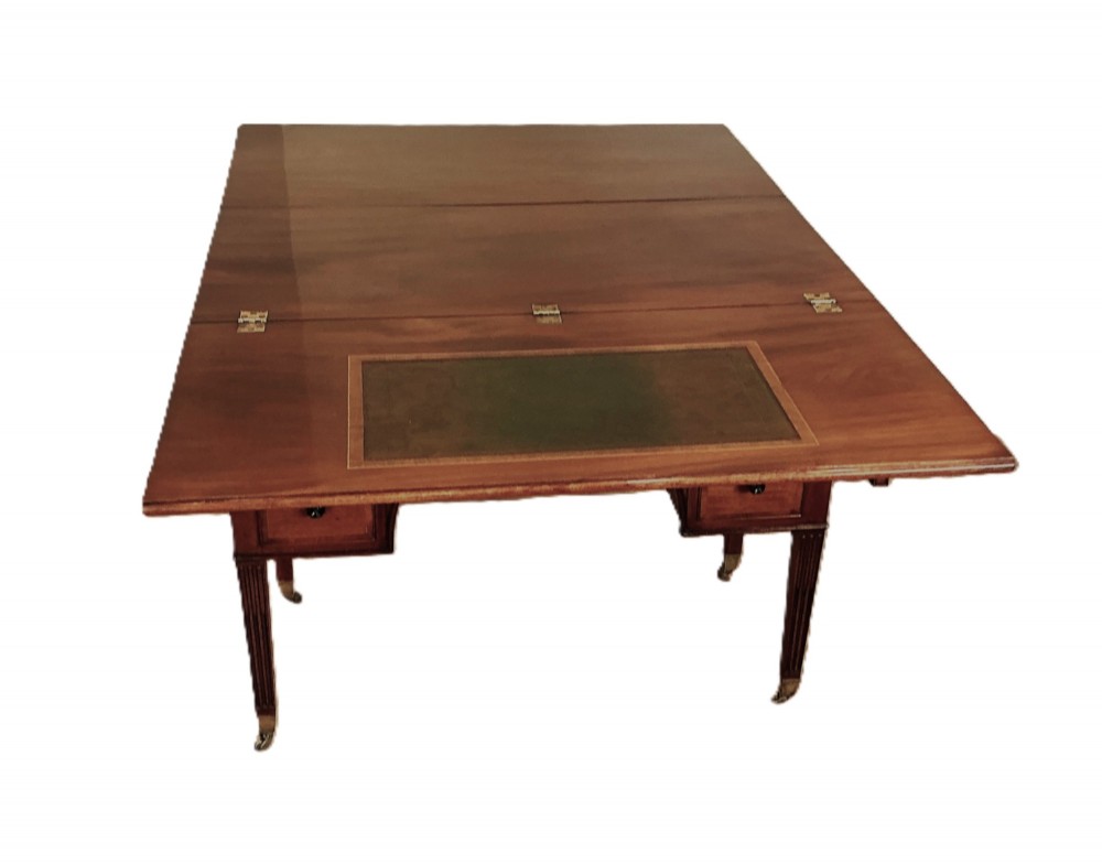 An antique George III mahogany library table featuring an arched central aperture, multiple drawers with dark turned knobs, and reeded tapered legs on brass castors. View II