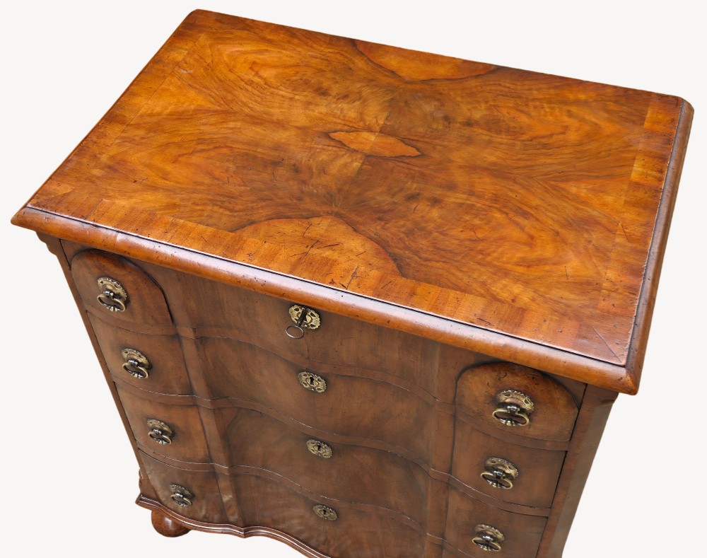 A Queen Anne style walnut chest of drawers featuring a serpentine front, brass drop handles, brass keyhole escutcheons, and bun feet. View III