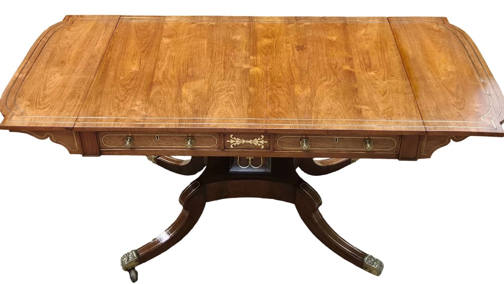 A Regency rosewood sofa table featuring hinged drop leaves, brass inlay decoration, two frieze drawers, and a quadripartite base on brass castors.