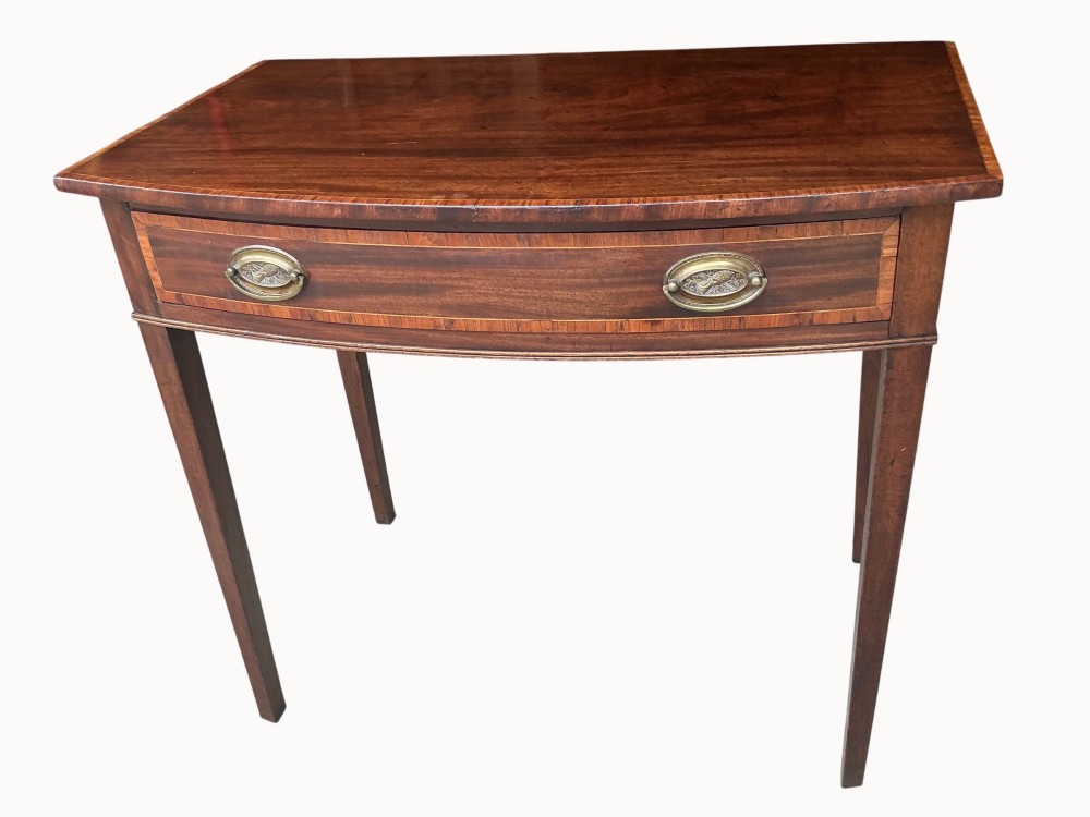 An antique mahogany Sheraton side table featuring a curved bow front, single crossbanded drawer with oval brass handles, and four square tapered legs.