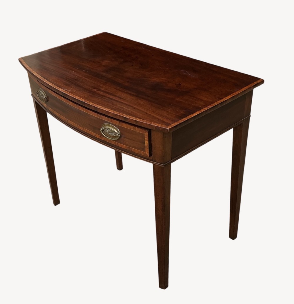 An antique mahogany Sheraton side table featuring a curved bow front, single crossbanded drawer with oval brass handles, and four square tapered legs. View II