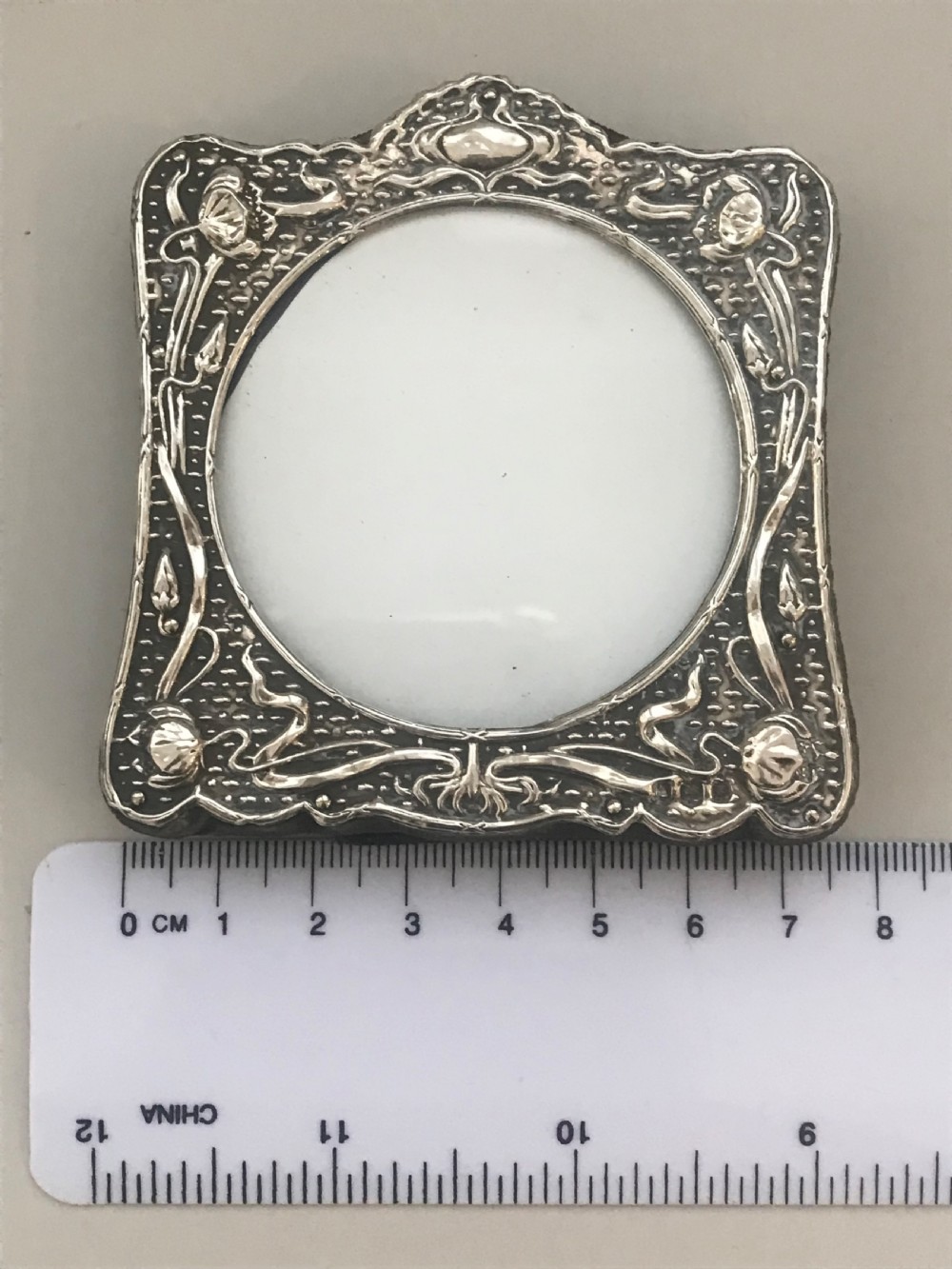 An Art Nouveau sterling silver photo frame featuring repousse floral motifs, a textured surface, and a central circular aperture. View II
