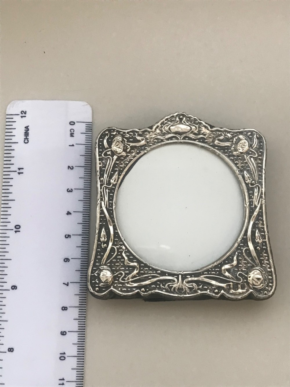 An Art Nouveau sterling silver photo frame featuring repousse floral motifs, a textured surface, and a central circular aperture. View III