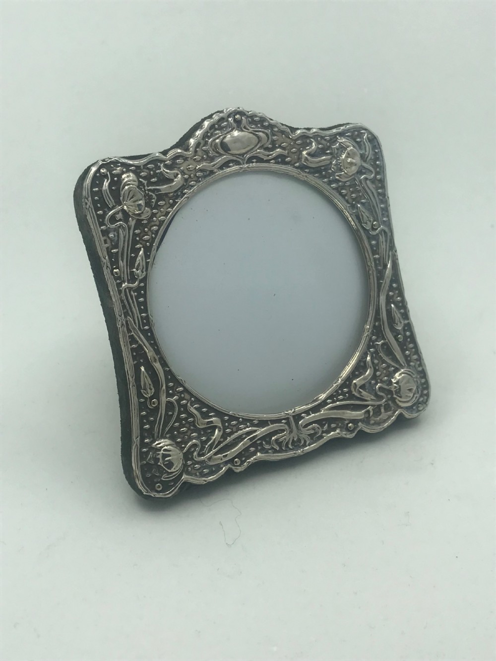 An Art Nouveau sterling silver photo frame featuring repousse floral motifs, a textured surface, and a central circular aperture. View VI