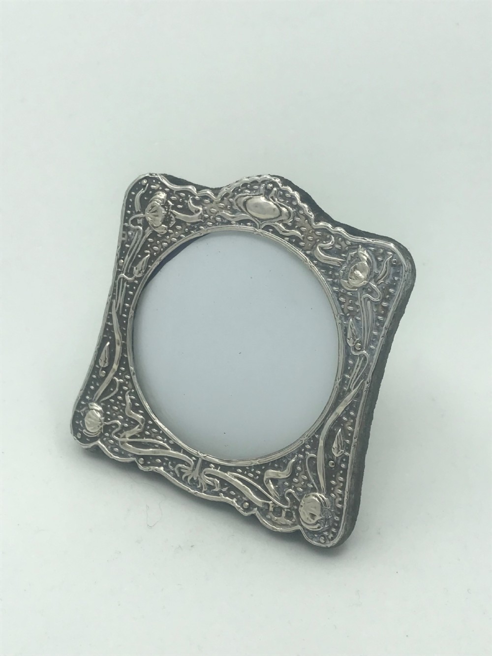 An Art Nouveau sterling silver photo frame featuring repousse floral motifs, a textured surface, and a central circular aperture. View VII