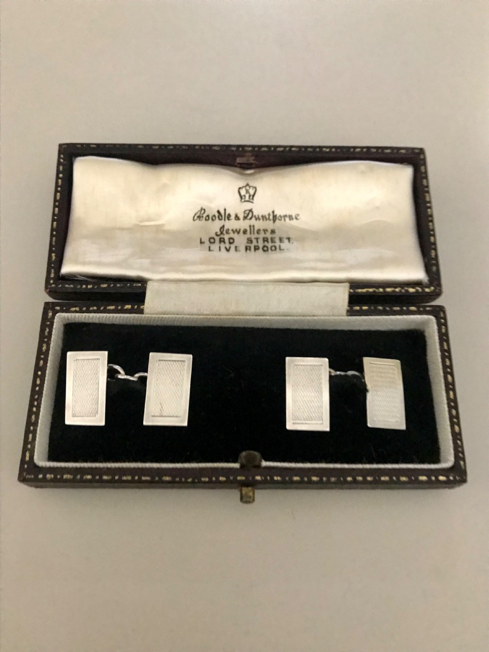 A pair of rectangular silver engine-turned cufflinks connected by chains, displayed inside an open fitted jewellers box.
